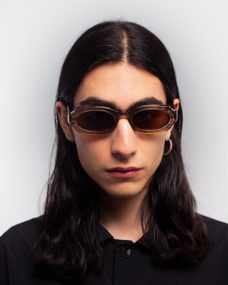 Model with long dark hair wearing translucent oval sunglasses and a black shirt against a neutral grey studio backdrop.