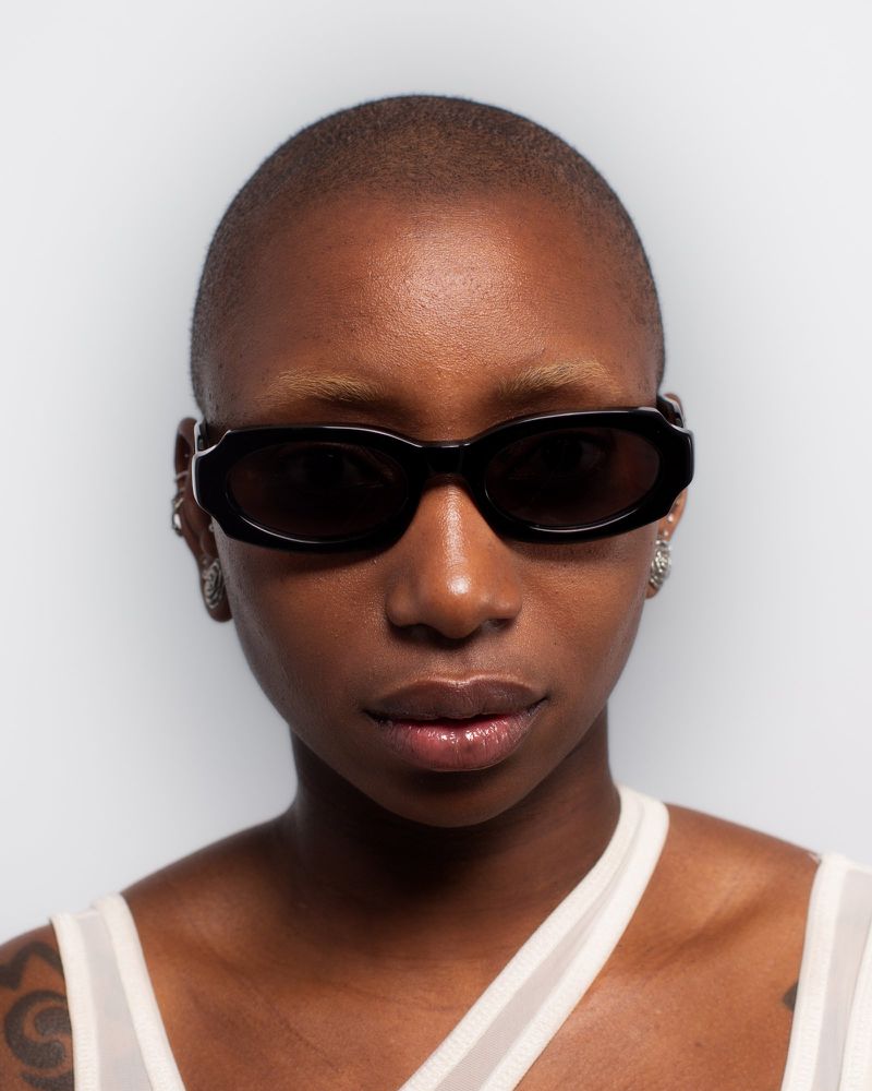 Close-up portrait of a Black woman with a shaved head wearing black rectangular sunglasses against a pale grey seamless background.