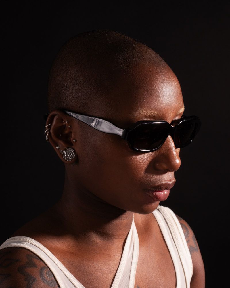 Close-up profile of a Black person with a shaved head wearing black sunglasses with silver arms against a black background.