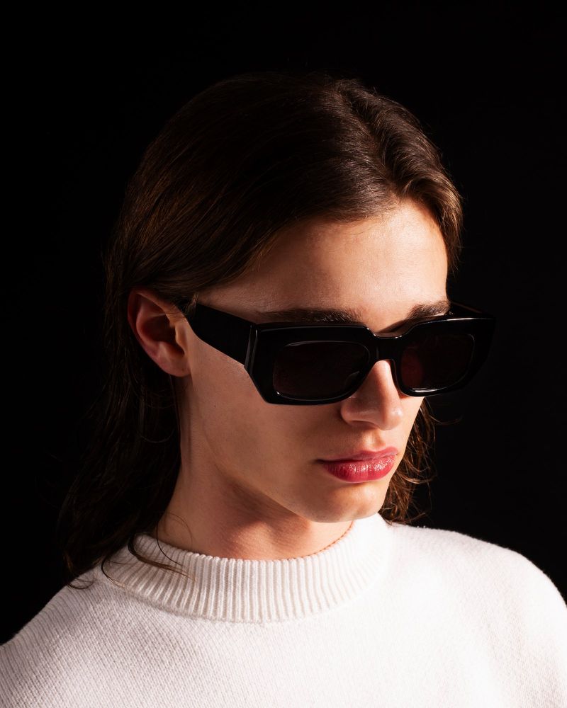 Model wearing chunky black rectangular sunglasses and a white crewneck knit sweater against a black studio background.