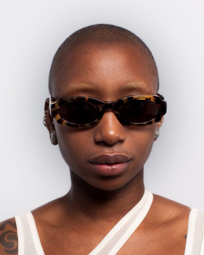 Close-up portrait of a woman with a shaved head wearing tortoise shell sunglasses against a plain white background.