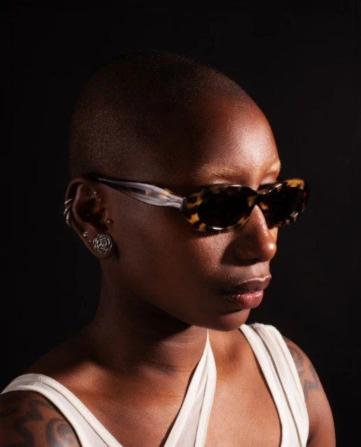 Portrait of an African American woman with a shaved head wearing sunglasses and jewelry against a black background.