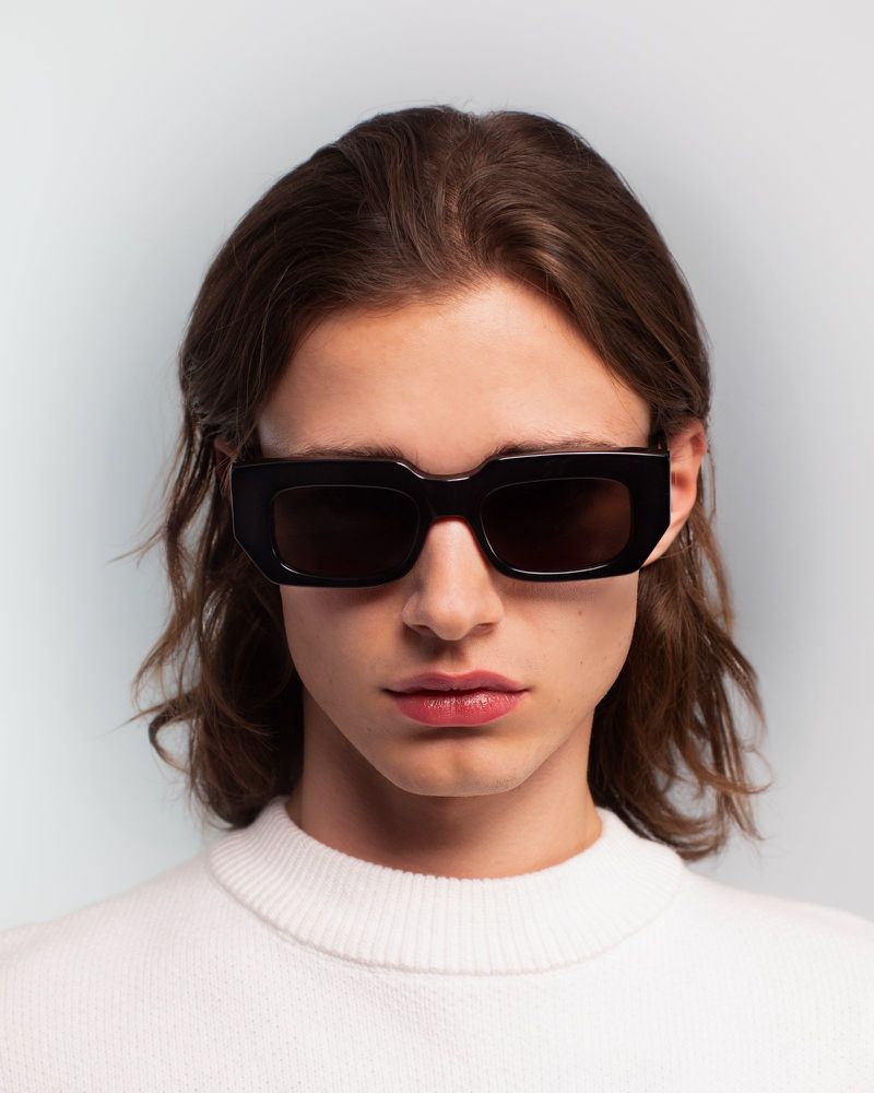 Person with long brown hair and wearing a white sweater and black sunglasses, posing in front of a solid white and grey backdrop.