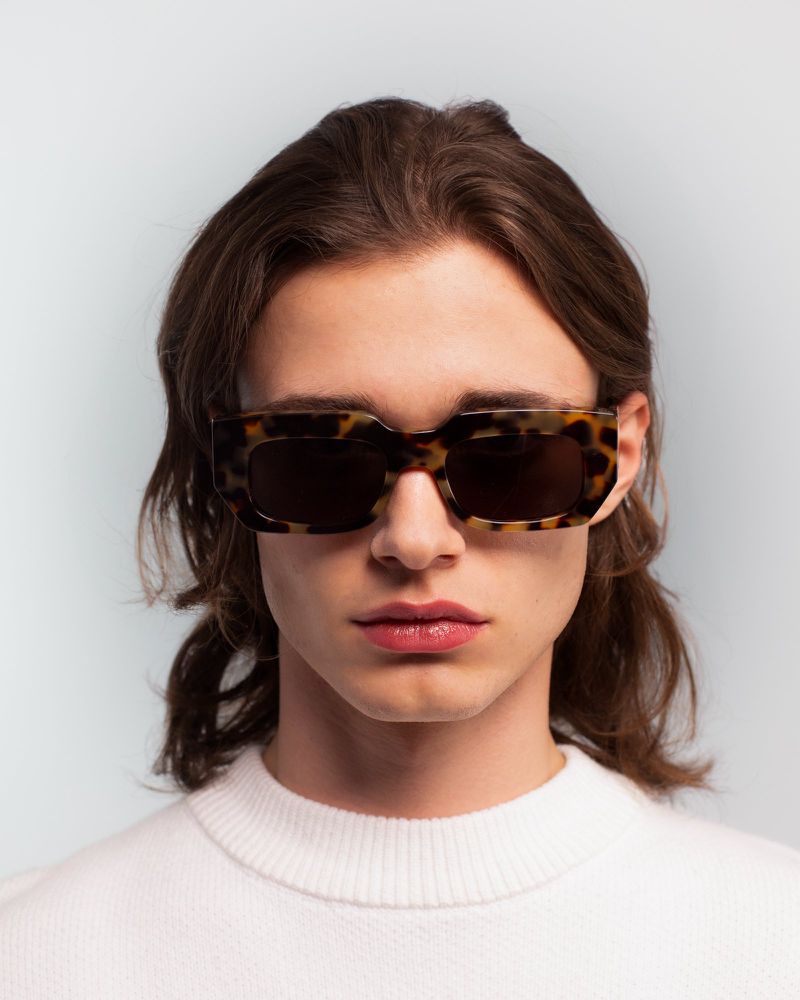 Close-up studio portrait of a young adult man wearing tortoise shell sunglasses and a white sweater against a light blue backdrop.
