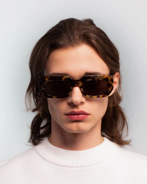 Close-up studio portrait of a young adult man wearing tortoise shell sunglasses and a white sweater against a light blue backdrop.