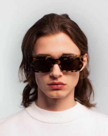 Close-up studio portrait of a young adult man wearing tortoise shell sunglasses and a white sweater against a light blue backdrop.