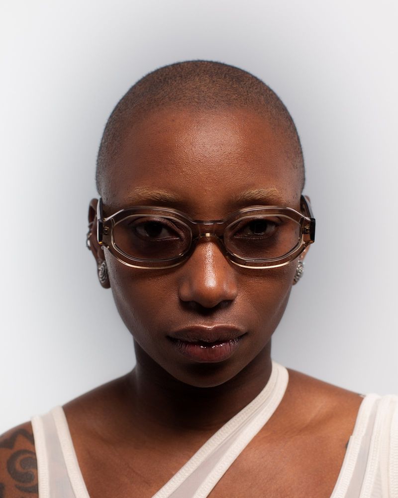 Close-up portrait of a bald African American woman wearing geometric glasses, large earrings, and a light-colored top against a white backdrop.