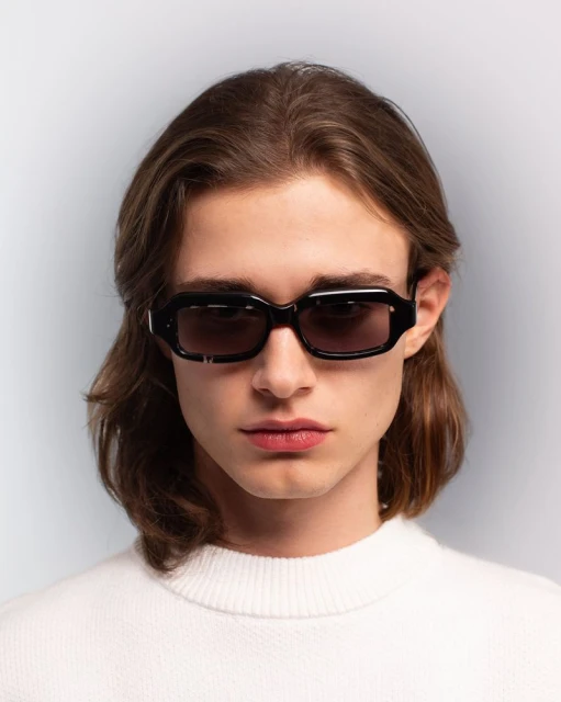 A portrait of a young adult male with brown hair wearing black sunglasses and a white sweater against a neutral gray background.