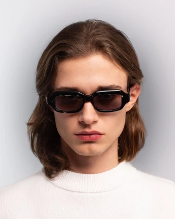 A portrait of a young adult male with brown hair wearing black sunglasses and a white sweater against a neutral gray background.