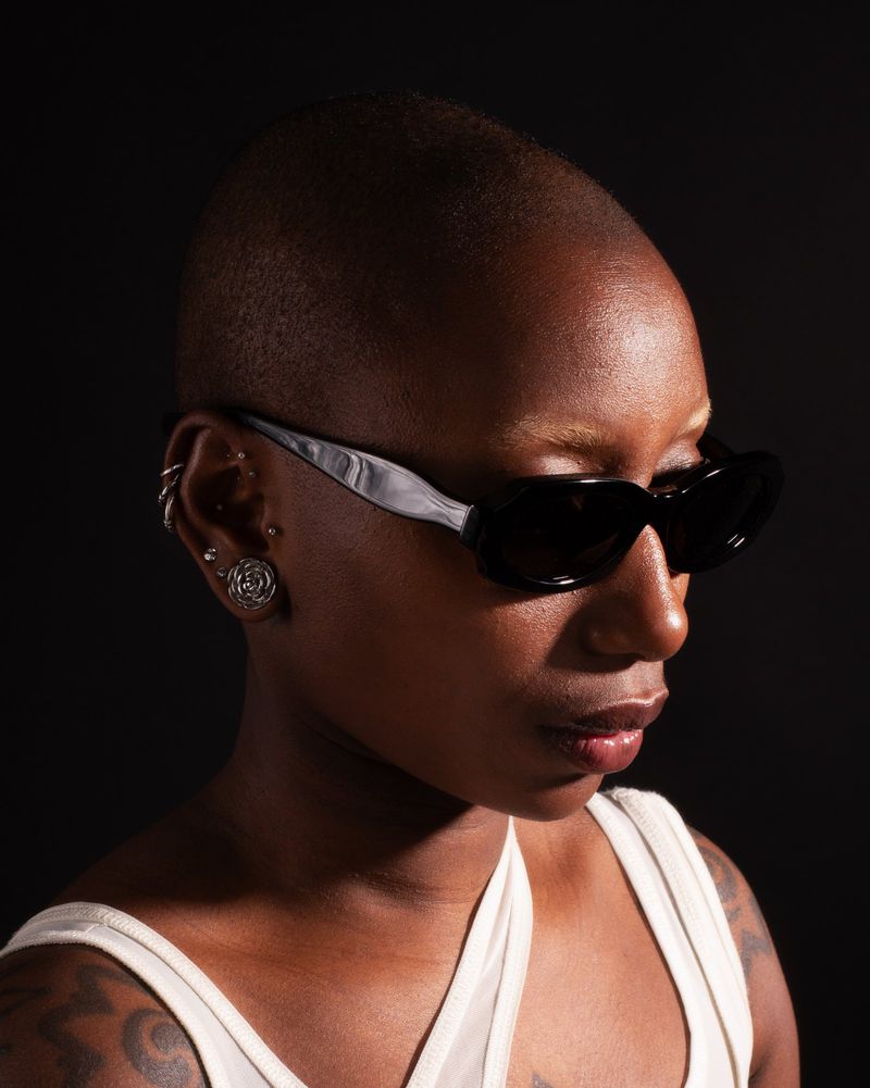 Profile portrait of a Black woman with a shaved head wearing sunglasses and a white tank top against a dark background.