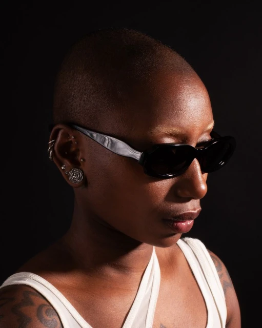 Profile portrait of a Black woman with a shaved head wearing sunglasses and a white tank top against a dark background.