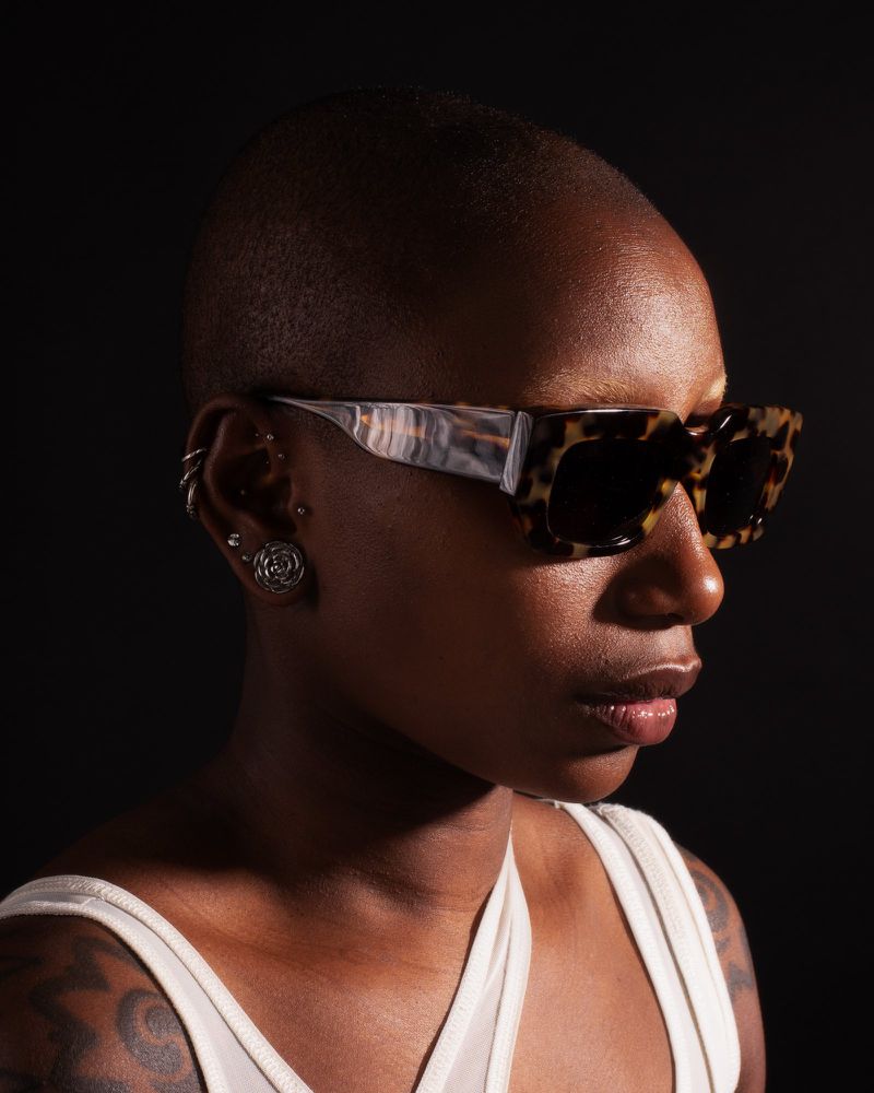 Adult female model poses in a sleeveless top and sunglasses against a dark backdrop, showcasing fashion and beauty.