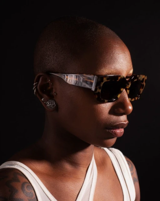 Adult female model poses in a sleeveless top and sunglasses against a dark backdrop, showcasing fashion and beauty.