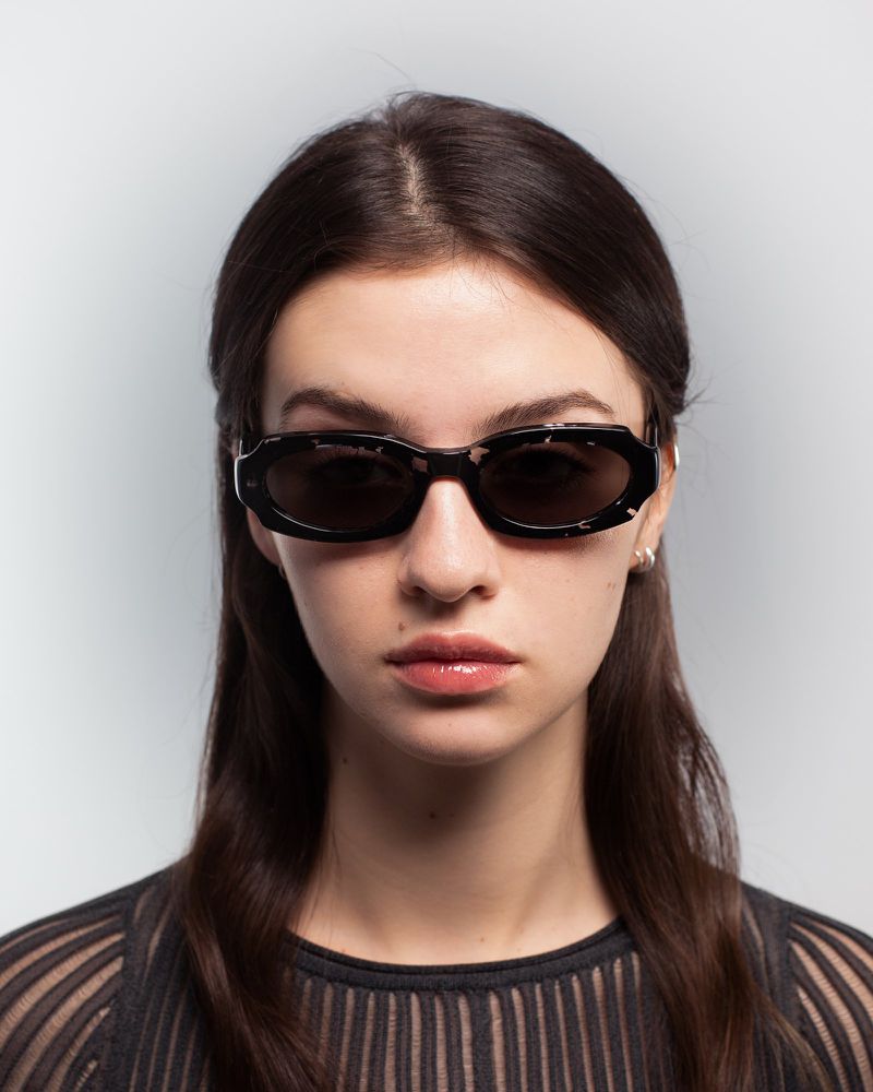 A woman with long brown hair poses in a studio wearing black-rimmed sunglasses, with a grey background and subtle studio lighting.