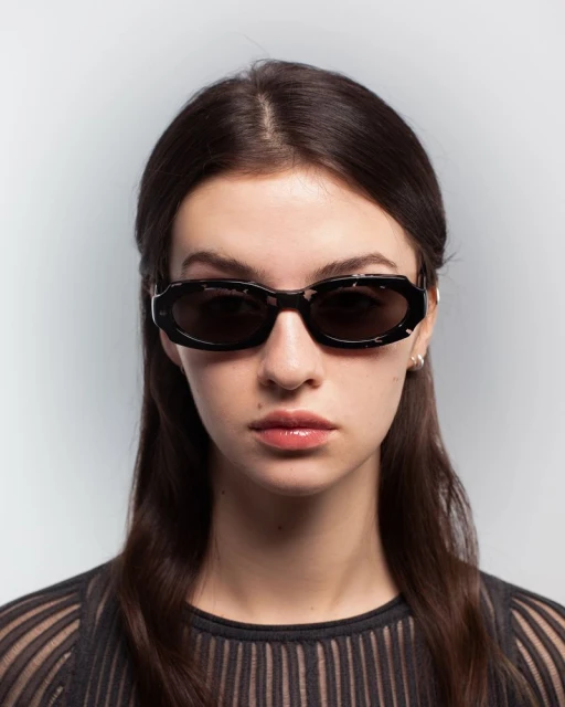A woman with long brown hair poses in a studio wearing black-rimmed sunglasses, with a grey background and subtle studio lighting.