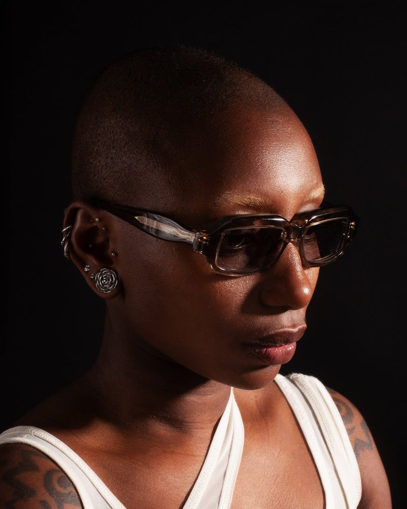 A portrait of a dark-skinned person with short hair wearing sunglasses, shot in a photo studio with a dark background and studio lighting.
