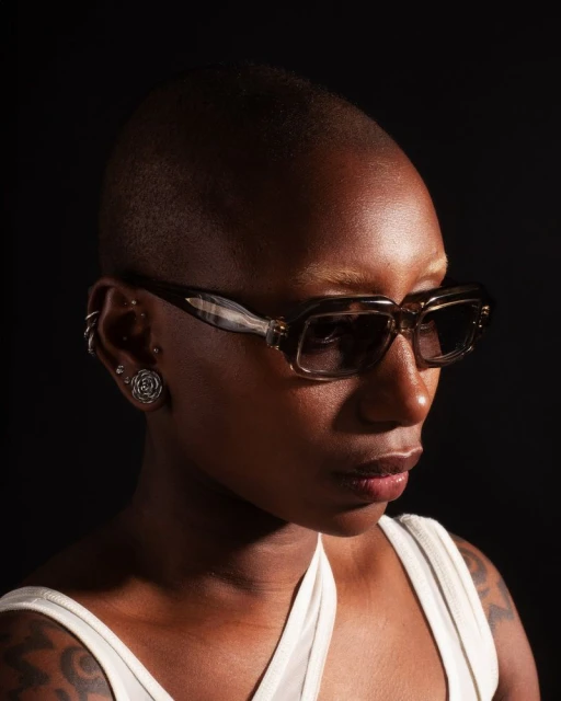 A portrait of a dark-skinned person with short hair wearing sunglasses, shot in a photo studio with a dark background and studio lighting.