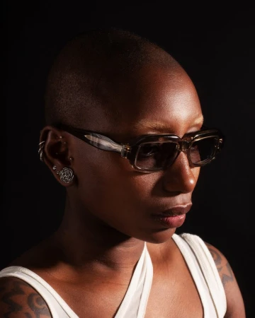 A portrait of a dark-skinned person with short hair wearing sunglasses, shot in a photo studio with a dark background and studio lighting.