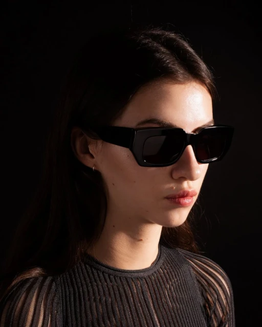 Close-up of a woman with dark hair wearing large, angular black sunglasses, set against a dark background with light highlighting her face.