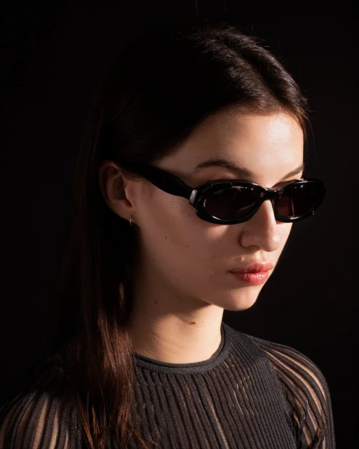 Portrait of a young adult woman wearing oval black sunglasses against a dark background, with shallow depth of field and soft lighting.
