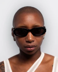 Close-up portrait of a Black woman with a shaved head wearing black rectangular sunglasses against a pale grey seamless background.