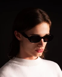 Model wearing chunky black sunglasses and a white crewneck knit sweater against a dark black background.
