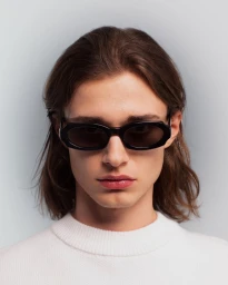 Close-up portrait of a man wearing black sunglasses and a white sweater against a pale background