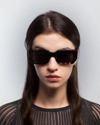 Close-up portrait of a woman wearing dark tortoiseshell sunglasses and a black ribbed top against a plain light grey background.