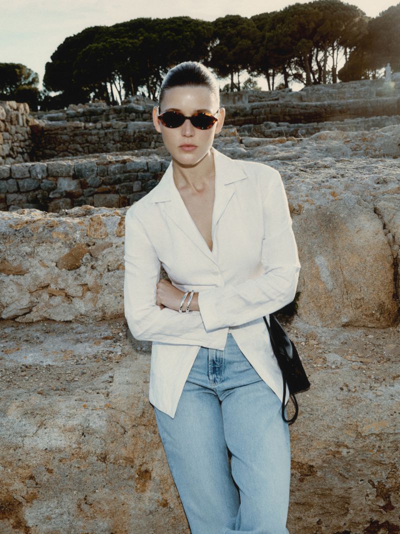 Fashionable adult woman in sunglasses poses in a white blazer and jeans against a stone wall with trees in the background.