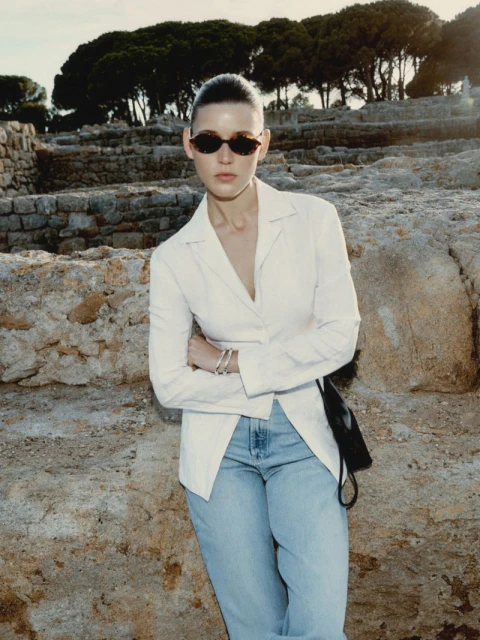 Fashionable adult woman in sunglasses poses in a white blazer and jeans against a stone wall with trees in the background.