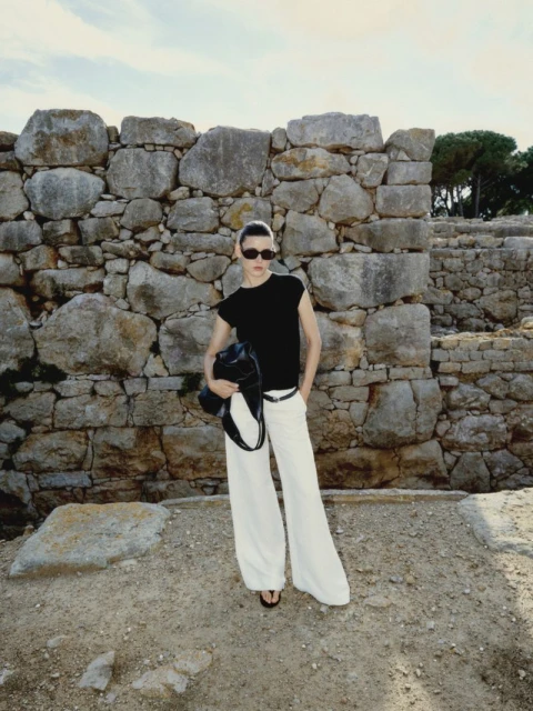 An adult woman wearing a black shirt and white pants stands near a stone wall holding a black purse in an outdoor environment.