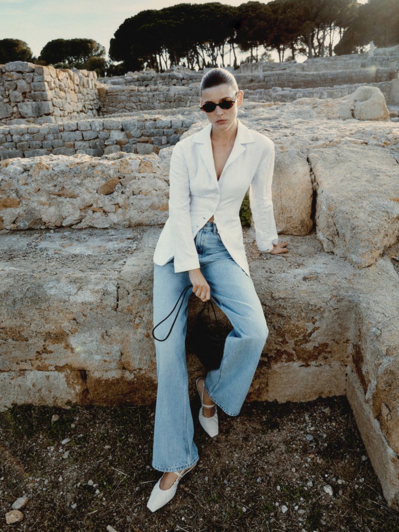 Woman in casual fashion posing against ancient stone ruins in outdoor setting with white linen jacket, blue jeans, black purse, and white shoes.