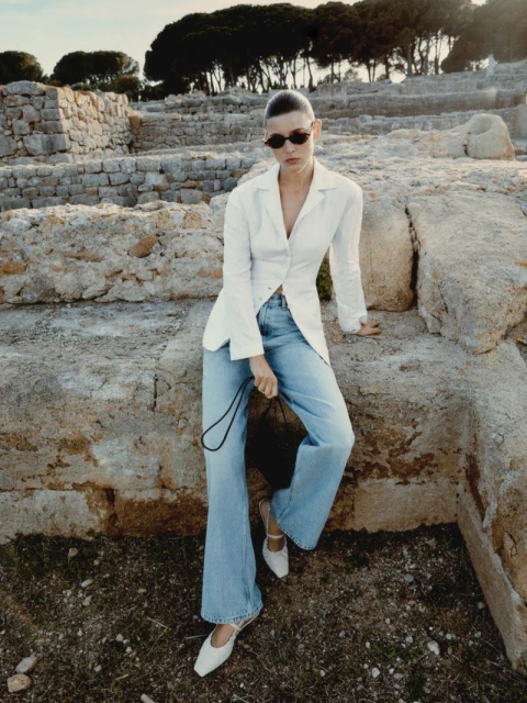 Woman in casual fashion posing against ancient stone ruins in outdoor setting with white linen jacket, blue jeans, black purse, and white shoes.