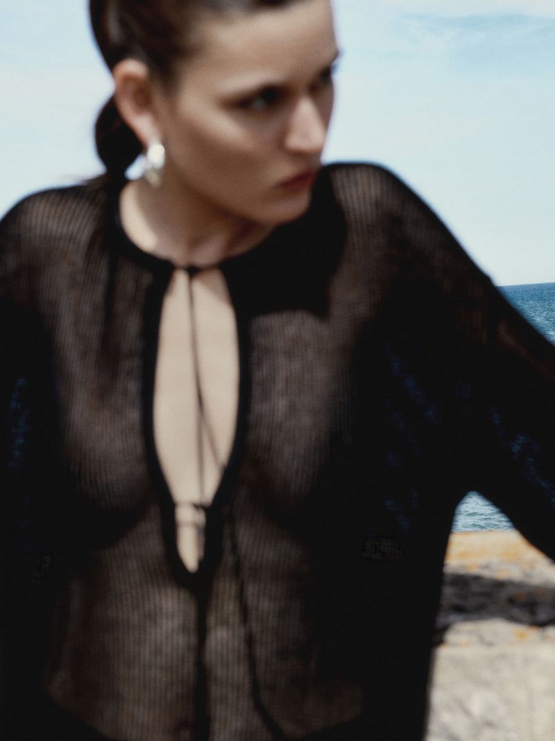 A woman gazes to the left, wearing a black mesh top with a keyhole neckline, against a backdrop of blue sky and ocean waters.