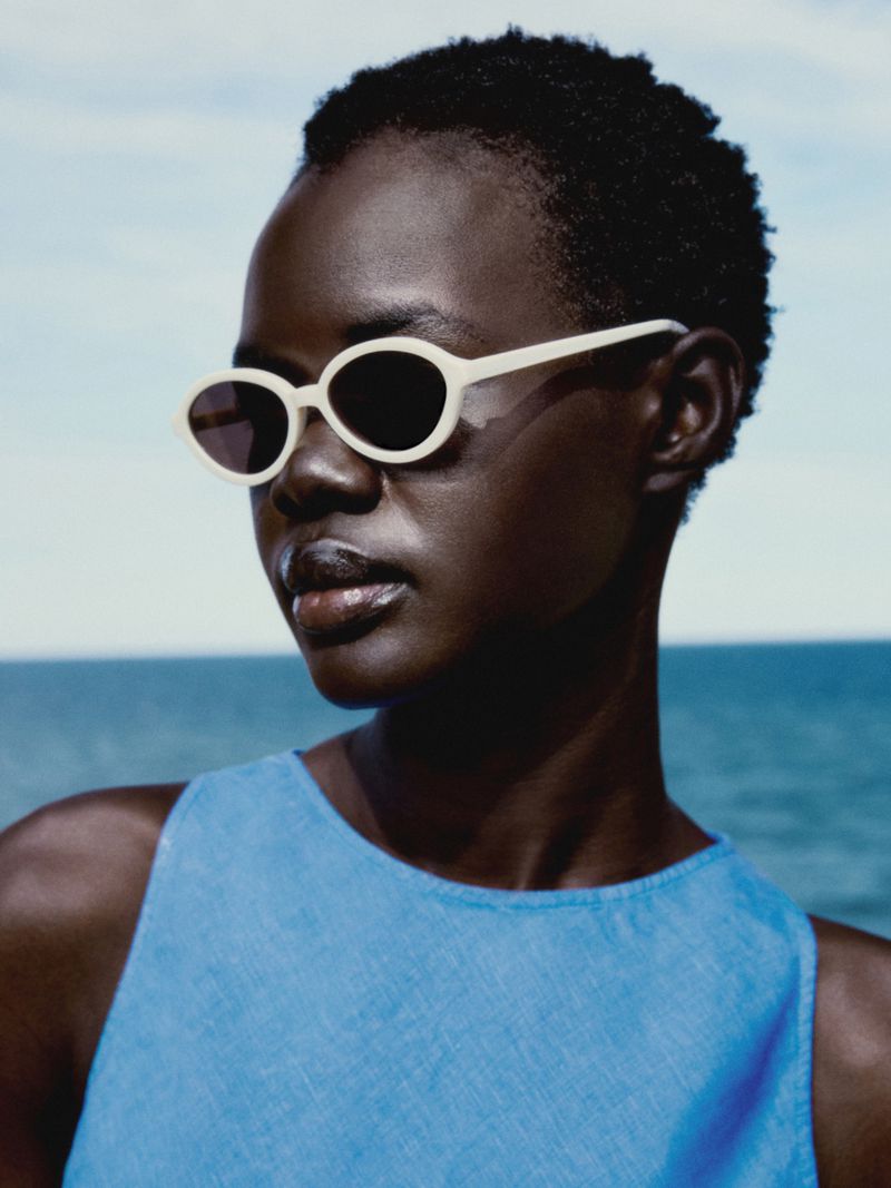 Close-up of a Black woman in a blue tank top and sunglasses against a bright sky and ocean backdrop on a sunny day.