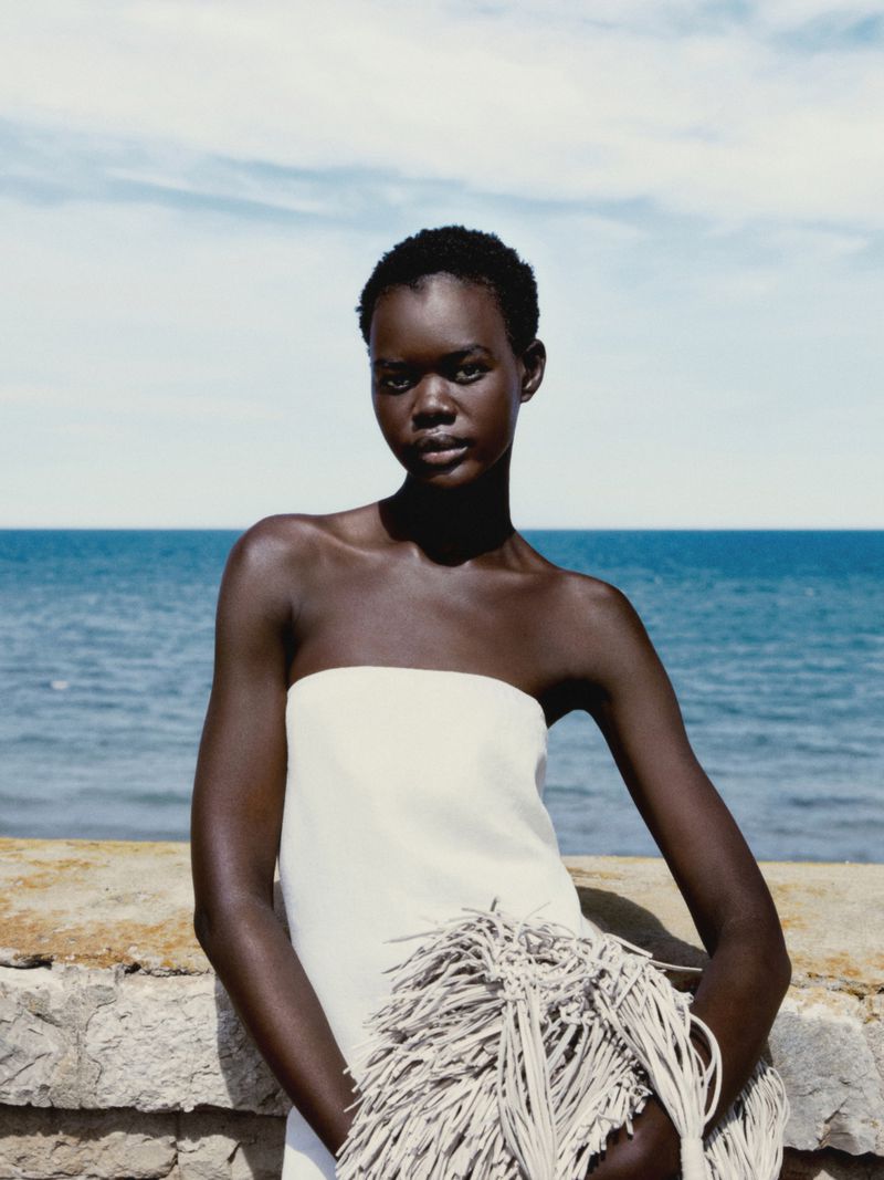 Model Kuany Atem in a white strapless dress holding a textured fringe bag against a blue ocean backdrop for Massimo Dutti.