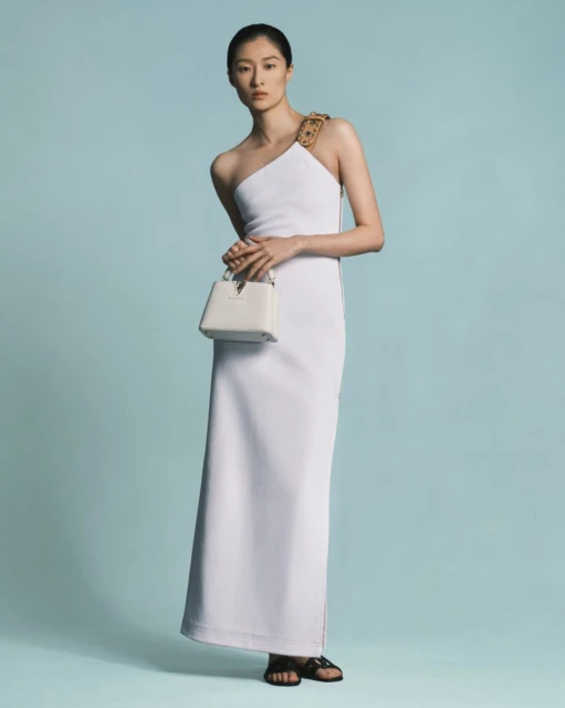 Elegant Asian woman posing in a white one-shoulder dress with a small white handbag against a light blue backdrop in a studio setting.