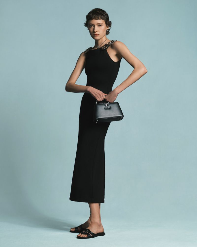 Full-length shot of an adult female posing in a black midi dress, black sandals, and holding a black top-handle handbag in a studio.