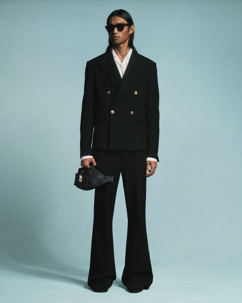 Model Ridzman Zidaine wearing a black double-breasted suit with gold buttons and flared trousers against a pale blue backdrop.