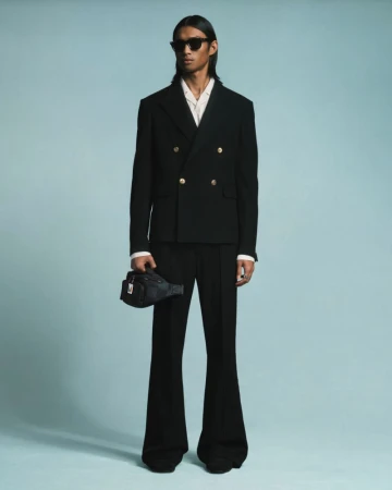 Model Ridzman Zidaine wearing a black double-breasted suit with gold buttons and flared trousers against a pale blue backdrop.