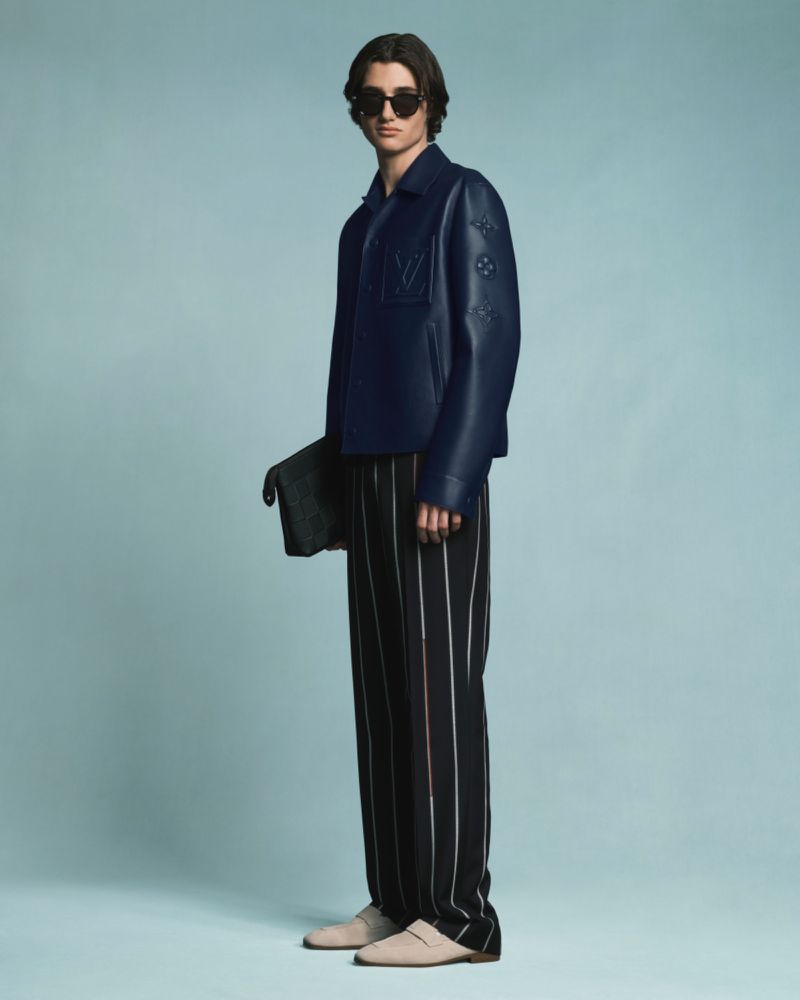 A person standing in a blue studio wearing black and white striped pants, a dark blue jacket, sunglasses, and carrying a clutch.