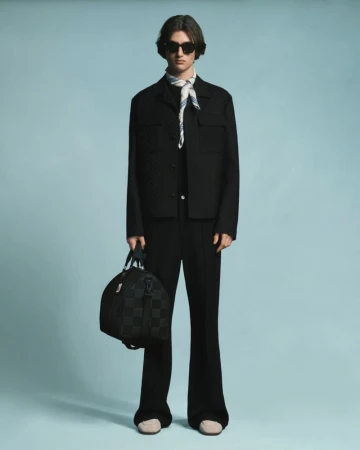 A full studio shot of a male model in a black suit, sunglasses, and a patterned scarf, holding a dark checkered duffel bag.