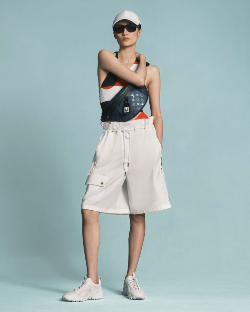 Full-length shot of a slender woman wearing a white cap, sunglasses, a patterned top, white shorts, and white sneakers, posing against a light blue backdrop.