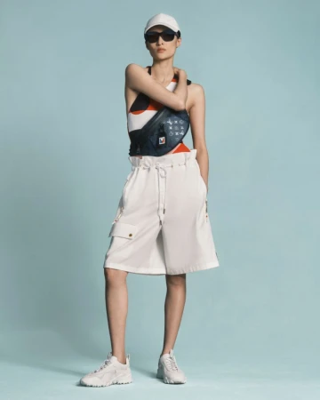 Full-length shot of a slender woman wearing a white cap, sunglasses, a patterned top, white shorts, and white sneakers, posing against a light blue backdrop.