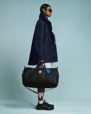 Side profile studio shot of a male model with sunglasses, holding a large travel bag, wearing a blue jacket with socks and sneakers.