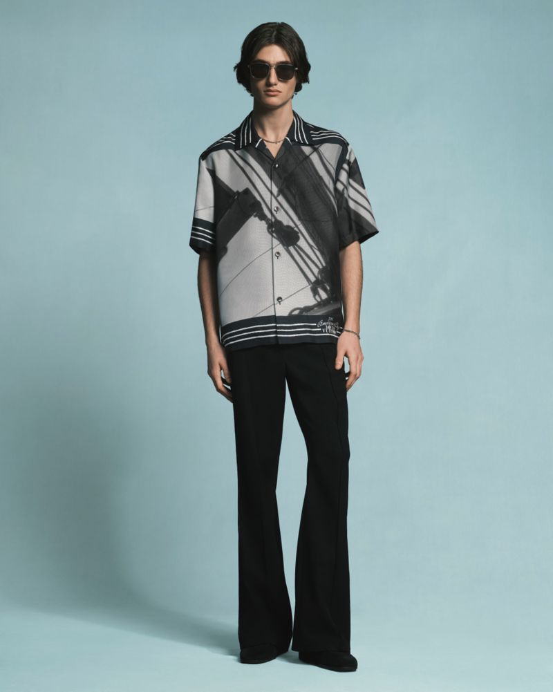 A young adult man stands in a photo studio wearing a patterned shirt, black pants, and sunglasses against a light blue background.