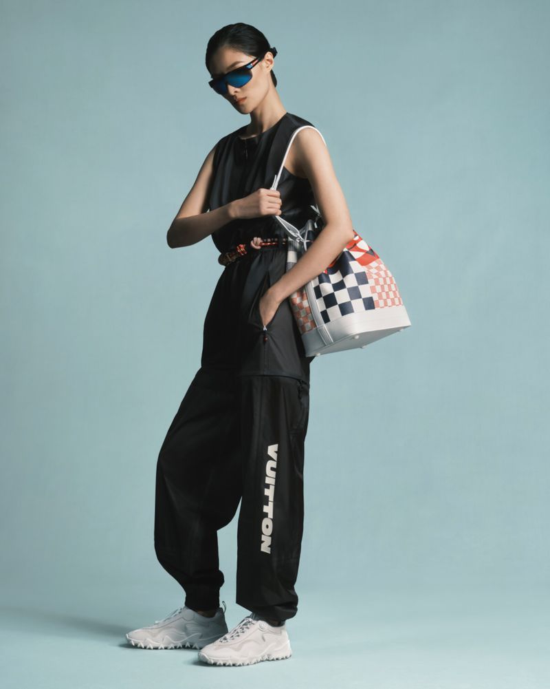 Woman in black sleeveless jumpsuit, sunglasses, and white sneakers poses with a checkered bucket bag against a light blue backdrop.