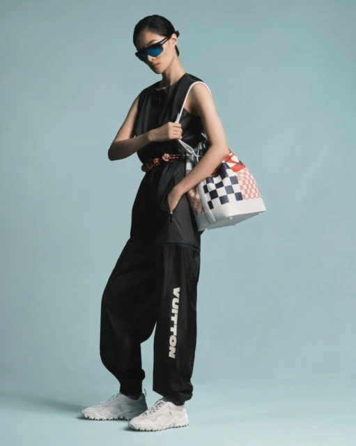 Woman in black sleeveless jumpsuit, sunglasses, and white sneakers poses with a checkered bucket bag against a light blue backdrop.