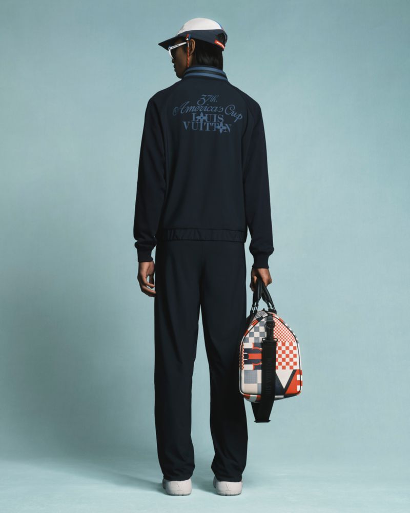A person wearing a navy blue tracksuit and a baseball cap, viewed from behind, holding a multicolored duffel bag against a light blue background.