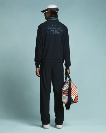 A person wearing a navy blue tracksuit and a baseball cap, viewed from behind, holding a multicolored duffel bag against a light blue background.